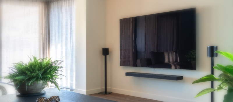 woonkamer met links witte gordijnen en rechts aan de muur een televisie inclusief soundbar en twee losse speakers op speaker standaarden