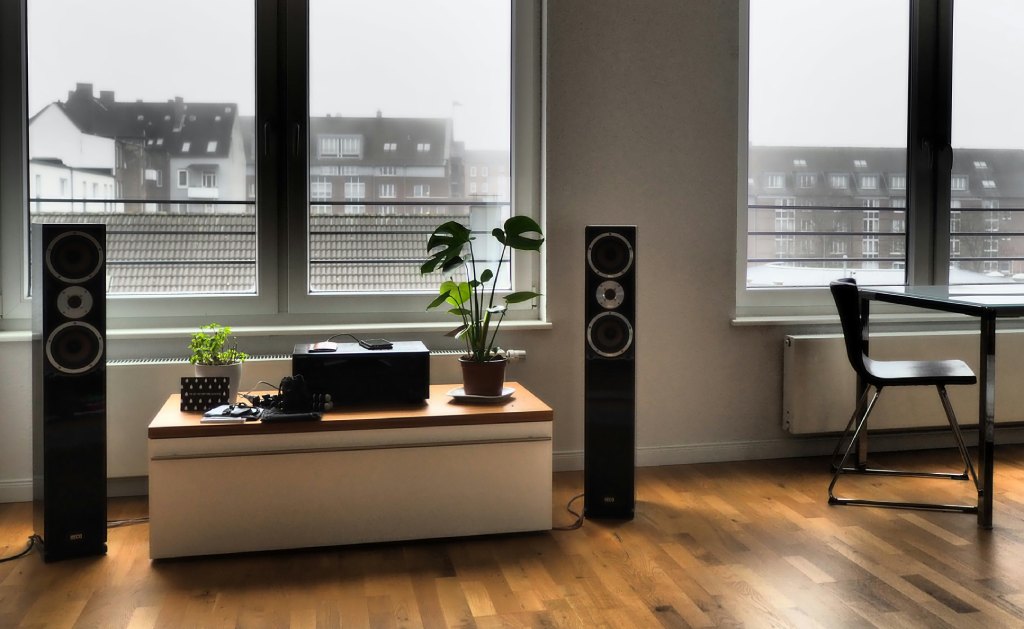 Een moderne woonkamer met grote vloerstaande speakers, een lage kast met planten en uitzicht op een stad.