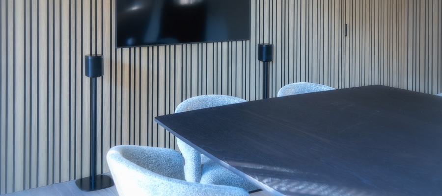 modern ingerichte kantoorruimte met een vergadertafel omringt met witte stoelen. Aan de muur hangt een televisiescherm met ernaast twee Vebos luidspreker statieven