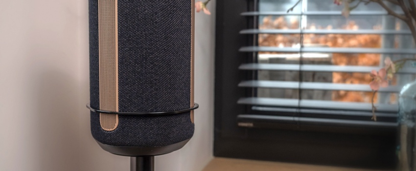 Close-up van een zwarte speaker op een Vebos standaard met een beugel, geplaatst in een modern interieur bij een raam met jaloezieën.